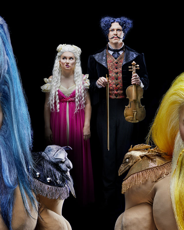Fantasilignende figurer med blå og gule parykker, foran en kvinne og en ann med fiolin i bakgrunnen