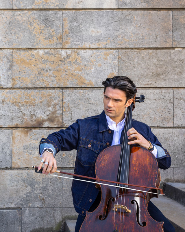 Mann sitter foran murvegg og spiller cello